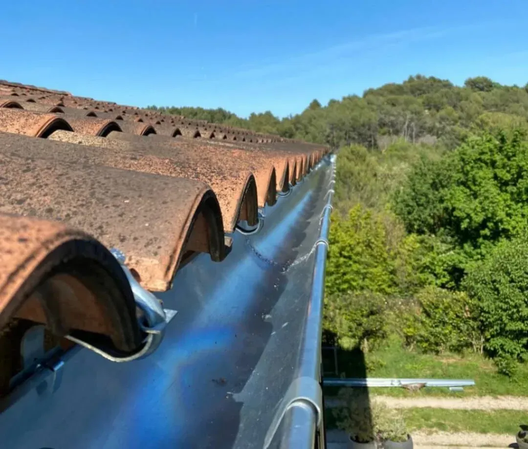 Gouttière en zinc – Installation sur toiture en Provence Gouttière en zinc fixée sous rangée de tuiles canal, ciel bleu et végétation en arrière-plan